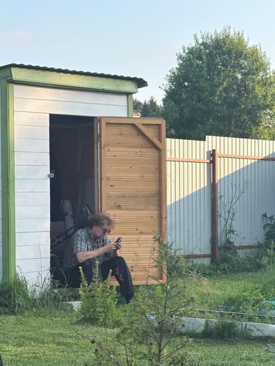 A young Caucasian man with curly blond hair and glasses is sitting in the open doorway of a white wooden shed with a green trim. He is wearing a plaid shirt and dark pants. He is looking at and holding a dark-colored smartphone in his hands, which he appears to be using. The shed's wooden doors are open. Inside the shed, a lawnmower and some bags are visible. The shed is situated in a grassy yard. A light-colored corrugated metal fence runs along the right side of the yard, with a wooden fence to the left of the shed. Trees are visible in the background. The scene appears to be in a rural or suburban area, potentially the city of Lyady, Russia, given the context. The lighting suggests it is daytime, possibly late afternoon due to the shadows.