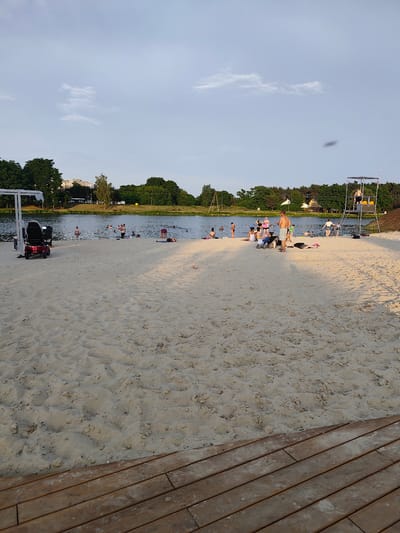 The image depicts a sunny day at a beach located in Stryków, Poland. A sandy beach extends into the foreground, with a wooden boardwalk in the lower right corner. In the middle ground, a body of water, likely a lake or river, is visible with numerous people swimming and wading. Several individuals are also present on the beach, some sunbathing, others interacting. To the left, a red mobility scooter is parked near a white gazebo structure. In the background, a line of trees and buildings forms the backdrop, with a tall lifeguard tower positioned on the right side of the beach. The sky is a pale blue with some scattered clouds.
