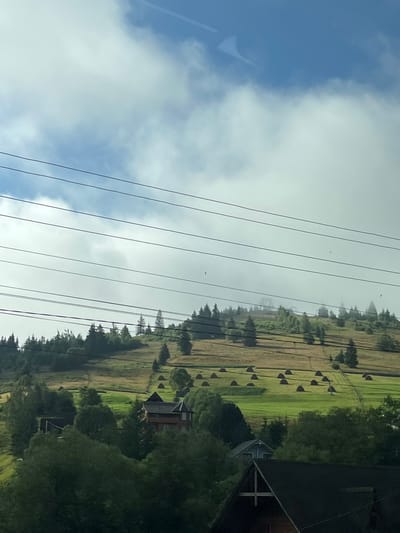 The image depicts a rural landscape in Yablunytsia, Ukraine, characterized by rolling green hills dotted with haystacks and trees. Several power lines stretch across the frame, cutting through the sky. A prominent wooden building with a distinctive roofline is visible on the hillside, and other smaller structures are scattered throughout the scene. The sky is a mix of blue and white clouds, with a hint of haze or fog clinging to the distant hills. The overall impression is one of a tranquil, natural setting with signs of human habitation.
