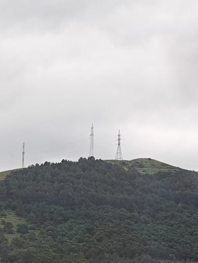 This media file depicts a cloudy day in Bazum, Armenia, with a landscape dominated by a forested hill. Three tall, lattice-style electricity pylons are visible on the crest of the hill, marking the passage of power lines across the terrain. The hill itself is covered in dense, dark green foliage, likely pine or fir trees, with patches of lighter green grassland interspersed. The sky is overcast with a uniform grey cloud cover, suggesting a cool or damp atmosphere. No people or text are visible in the image. The scene evokes a sense of rural remoteness and the utilitarian presence of infrastructure within a natural setting. The time of day appears to be daytime, given the general illumination, but the cloudy conditions prevent distinct shadows or direct sunlight.