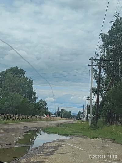 The image depicts a rural road in Balagansk, Russia, on July 11, 2026, at 13:53. The road is unpaved and has large puddles from recent rain. On the left side of the road, there is a decorative fence and lush green trees. On the right, utility poles with numerous wires stretch along the road, interspersed with trees. In the distance, more trees and buildings can be seen, with a cloudy sky overhead.