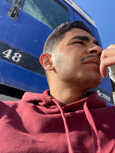 A man with dark hair and a maroon hoodie poses in front of a blue truck. The truck's front is visible, displaying the number "48" and part of the word "ACTROS" in white lettering. The man's face is partially obscured by his fingers, and his eyes are closed. The background shows a clear, light blue sky. The image appears to be taken from a low angle.