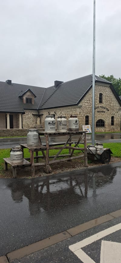 In Kaali, Estonia, an overcast sky looms over a stone building with a dark, tiled roof. The building, identified by signage as "KAALI TRAHTER RESTORAN", features arched windows and a dormer window. In front of the building, a rustic wooden table is set up, displaying several large metal milk churns. Three churns are placed on the table, each with labels indicating "KAALI TRAHTER", "ANGLA", and "PANGA PANK". To the left, another churn sits on a wooden step, labeled "TURISM 'PIIMARING'". A smaller churn is placed on a lower wooden step and is labeled "TERE TULEMAST WELCOME ДОБРО ПОЖАЛОВАТЬ". A small, wheeled cart with two more churns is positioned to the right of the table. The ground is wet and reflective, indicating recent rain. The scene appears to be set up as a display or welcome area for visitors.