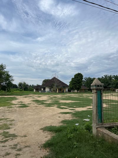 The image depicts an outdoor scene, likely in Landuh, Indonesia, under a partly cloudy sky. In the foreground, a dirt path transitions to a patchy lawn with scattered debris. To the right, a weathered concrete pillar with a green metal fence attached stands prominently, bordering the property.
In the middle ground, several dilapidated buildings with sloping roofs are visible, surrounded by lush green vegetation and trees. One building in particular, with a dark, pyramid-shaped roof and pale walls, appears to be the most substantial structure. The buildings seem abandoned or under construction, with some windows boarded up.
The background is dominated by a dense arrangement of trees, including palm trees, suggesting a tropical or subtropical climate. Overhead, thin, wispy clouds streak across a pale blue sky. Power lines are visible in the upper right corner of the frame. The overall impression is one of a neglected rural or semi-rural setting.
