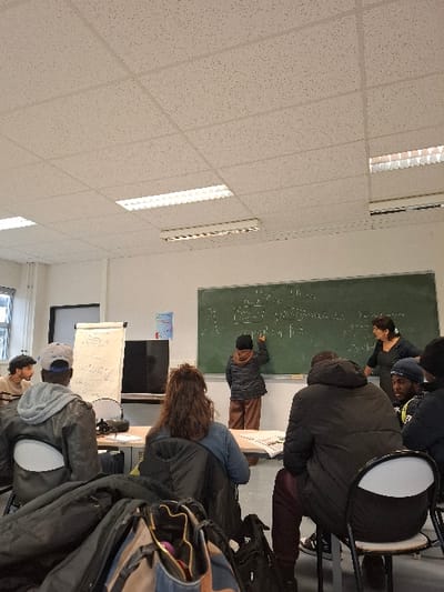 A classroom scene shows students around a chalkboard filled with equations. Some students are actively writing on the board, while others observe. The foreground features a person wearing a dark jacket, with bags and personal items arranged on a chair nearby. The lighting is bright, emphasizing the white ceiling tiles with recessed lighting. The classroom atmosphere suggests active learning and collaboration.