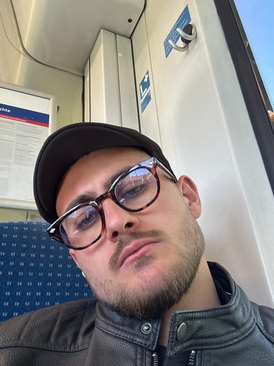 This image is a portrait-style shot of a man, likely a selfie, taken inside a train carriage. The original image is rotated 90 degrees clockwise; when viewed upright, the man's face fills a significant portion of the frame, looking directly towards the camera.
The subject is a man with short facial hair/stubble. He is wearing a black flat cap and a black leather jacket, visible on his right shoulder and arm. He has distinct tortoiseshell-framed glasses with visible reflections, suggesting light sources in front of him. His expression appears neutral and calm, with his eyes open.
The setting is the interior of a public transport vehicle, specifically a train, as indicated by the various details and the location context of Geldern, Germany. The background shows the train's internal structure: white/off-white ceiling panels on the left with a bright window area at the very top, suggesting daylight. A silver, double-pronged hook is visible on the ceiling panel, along with small blue stickers, one of which appears to have a cross symbol. On the right and below the man is blue patterned seat upholstery. A white information board or poster is partially visible below the man's chin.
Visible text on this information board includes "chte" (possibly part of a word like "Rechte" or "Geschichte") and "rsummis von Anschlußzügen," which translates to "delay of connecting trains." This text confirms the German context and indicates the poster likely pertains to travel information or passenger rights. The small blue stickers on the ceiling have illegible text but show graphic symbols.
The bright light from the top window suggests it is daytime. The scene captures a quiet moment of travel.