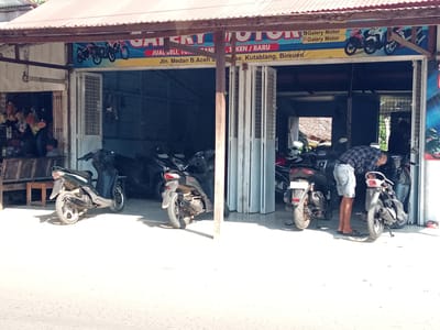 The image shows the exterior of "Galery Motor" in Blangpanjoe, Indonesia. The shop entrance is open, revealing several motorcycles parked inside. A man wearing a plaid shirt and denim shorts is tending to a motorcycle, bending down to inspect it. To the left, a green scooter is parked outside the shop. The shop's signage features images of motorcycles and text that appears to indicate the sale of new and used vehicles, along with an address. The environment is bright and sunny, suggesting daytime.