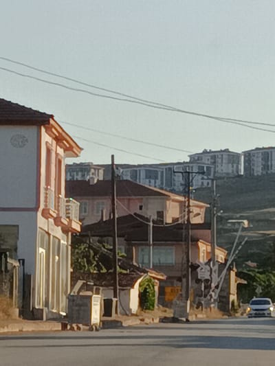This image depicts a street scene in Bahşılı Belediyesi, Turkey, under clear daylight conditions. The foreground features a paved road, with a sidewalk on the right. Several buildings line the street. The building on the far left is light-colored with orange trim and windows. Adjacent to it are other structures with terracotta roofs and varying facades. A large grey dumpster is visible on the sidewalk.
In the mid-ground, a series of multi-story apartment buildings are situated on a gentle slope. These buildings appear to be modern in design. A network of power lines crisscrosses the sky above the street and buildings.
In the background, the landscape rises, with more buildings visible on the hills. A white car is driving on the road in the right side of the frame, its headlights on. The lighting suggests it is daytime, likely late afternoon or early evening due to the warm glow on the buildings. There is no visible text in the image. The scene conveys a sense of everyday life in a semi-urban or suburban area.