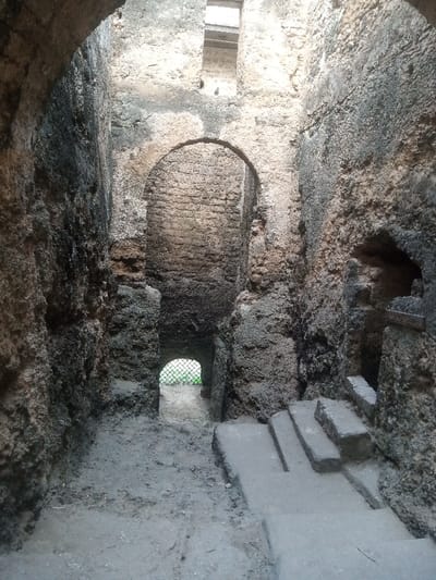 The image shows an ancient coral stone passage within Fort Jesus, located in the Makadara/Mji Wa Kale ward, Mombasa, Kenya. The high, rough-hewn stone walls display significant weathering, erosion, and dark biological staining. A prominent arched entryway is visible centrally in the mid-ground, leading further into the structure. At the passage's end, a smaller, lower arched opening reveals a metal grate and distant green vegetation. On the right, a set of uneven, rectangular stone steps ascends. A small rectangular wooden sign affixed to the right wall above the steps displays the word "SLAVERY" in faded white lettering. Natural light enters from a rectangular opening positioned high above and from the distant arched exit, creating varied illumination across the textured stone surfaces. No individuals are present.