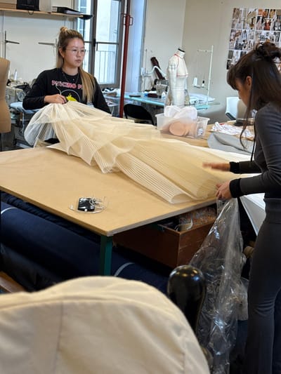 The image depicts an interior scene within a fashion or textile design workshop located in Arcueil, France. Two women are actively engaged in working on a large, off-white, intricately pleated fabric garment or textile piece laid flat on a light-colored wooden work table.
The woman on the left, of East Asian descent, has light brown hair tied in a bun and wears glasses. She is dressed in a black long-sleeve shirt with a yellow "PULCINO PI" graphic and is positioned behind the table, handling an upper section of the pleated fabric.
The woman on the right has dark hair tied up and is wearing a dark grey ribbed long-sleeve top and dark pants. She stands to the right of the table, using both hands to manipulate a lower section of the pleated fabric.
A black smartphone with white wired earbuds rests on the table surface, positioned between the two women and the fabric item.
In the background, behind the woman on the left, a window shows an indistinct building exterior. Nearby, a sewing machine and a mannequin or dress form are visible. Behind the woman on the right, another dress form marked with red lines, a thread stand, and other sewing equipment are present. The wall behind them is adorned with numerous pinned-up fabric swatches or design inspirations.
Underneath the work table, several rolls of dark blue fabric are stacked, and a wooden box containing various colored textile scraps or threads is partially visible. A clear plastic bag rests near the right woman's legs. The shoulder and upper back of a third individual, clad in a light-colored garment, are visible in the immediate foreground, out of focus.