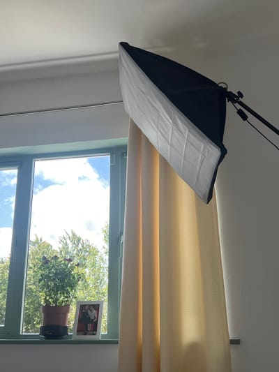 The image depicts an indoor scene, likely a room in a residential property located in Balbriggan, Ireland. The primary focus is on a window with a light blue-green frame, revealing a view of lush green trees and a bright blue sky with scattered white clouds. On the windowsill, there is a potted plant with small red flowers and a framed photograph of a couple embracing. To the right of the window, a beige curtain with a subtle dotted pattern hangs, partially obscuring the wall. Above and to the right of the window, a photographic softbox, a lighting device used in photography, is positioned on a stand. The softbox has a black outer casing and a white diffusion panel, angled downwards towards the scene. The overall impression is that of a home setting, possibly being prepared for or used in a photography session.