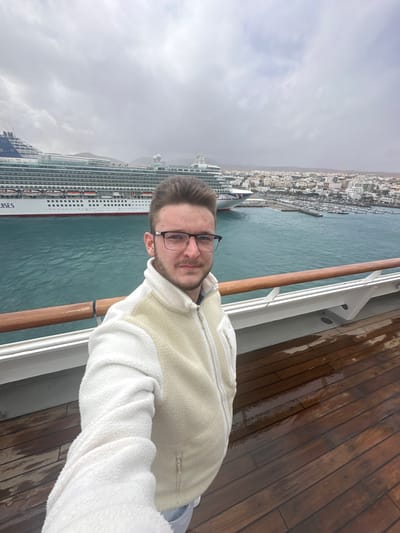A young adult male, wearing a cream-colored fleece vest over a white long-sleeved fleece, stands on the wet wooden deck of a ship and captures a selfie. He wears black-framed glasses and has short brown hair and light facial stubble. Behind him, turquoise water fills the midground, separated by a white railing with a wooden top. A large white cruise ship, identified by "P&O CRUISES" on its bow and multiple decks, is docked or moored in the port of Puerto del Rosario, Spain, positioned to the left. To the right of the cruise ship, a marina crowded with smaller boats and yachts is visible. The city of Puerto del Rosario, characterized by numerous low-rise, light-colored buildings, extends along the coastline into the background. Arid hills rise in the far distance, beneath an overcast sky with extensive grey cloud cover.