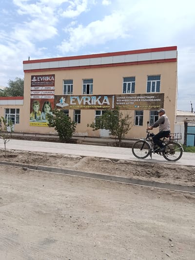 The image shows a street scene in Kungrad, Uzbekistan, featuring a two-story building with signage for a children's educational center. The building is painted beige with a red and white striped trim at the top. On the left side of the building, a banner displays "EVRIKA" along with text in Russian, advertising the "CHILDREN'S EDUCATIONAL CENTER OF SCHOOL AND EARLY AGE." Below this, there are smaller sections listing subjects such as "PREPARATION FOR SCHOOL," "RUSSIAN LANGUAGE," "MENTAL ARITHMETIC," "MATH," "ENGLISH LANGUAGE," and "SPEECH DEVELOPMENT." A large image of two smiling children is also part of this banner. Further to the right, another banner reads "EVRIKA" with the tagline "THE BEST INVESTMENT IS AN INVESTMENT IN THE EDUCATION OF YOUR CHILDREN," also in Russian, followed by a phone number and an Instagram handle "@evrika_kungrad."
In front of the building, a man wearing a cap and a grey vest over a long-sleeved shirt is riding a bicycle. The bicycle has a basket on the back and appears to be a moped or motorized bicycle. He is traveling from left to right across the frame.
The environment includes a paved sidewalk in front of the building, with a dirt area and a curb bordering it. The street in the foreground is unpaved and appears dusty. Trees are visible along the sidewalk. The sky is partly cloudy. The overall impression is an urban street on a bright day, with evidence of ongoing development or maintenance in the foreground.