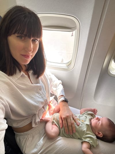 A medium shot angled from the side shows a woman and an infant inside an airplane. The woman is lying down in a white button-down shirt, with her dark hair spread across the fabric and her face turned towards the camera. She has dark hair with bangs and fair skin. The infant is lying on top of the woman, with their head near the top of the frame. They are wearing a light green onesie with a giraffe graphic. A hand wearing a watch and wedding ring is holding the infant. The airplane window is visible in the upper left, with the outside light suggesting daytime. The scene is presumably inside a commercial airplane, but there is no further information about the location of Cinisi, Italy.