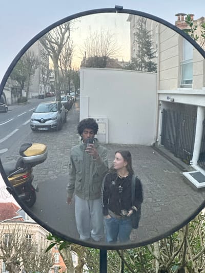 The image is a reflection captured in a circular, convex security mirror positioned outdoors, depicting two individuals and a street scene in Le Perreux-sur-Marne, France.
In the foreground of the reflection, a male figure with dark, curly hair is positioned on the left, wearing a green multi-pocket jacket over a light-colored hooded garment and light grey sweatpants. He holds a smartphone elevated, seemingly taking a photograph of himself and the second individual. To his right, a female figure with shoulder-length light brown hair is visible, dressed in a dark long-sleeved top with white stripes on the sleeves, light blue jeans, and multiple necklaces including one with a yellow pendant. She looks towards the male and the smartphone, with her left hand in her jeans pocket.
The background, reflected in the mirror, shows a paved street with a white road marking along the left side. Several vehicles are parked, including a silver Renault Clio (French license plate BY-845-RC) and a yellow-topped scooter on the sidewalk. Bare deciduous trees line the street. On the right side of the reflection, a light-colored building features a dark, gridded garage entrance and a window with a white roll-down shutter. The sky is overcast, providing diffused ambient lighting to the scene. Green foliage partially obscures the lower edge of the mirror's frame.