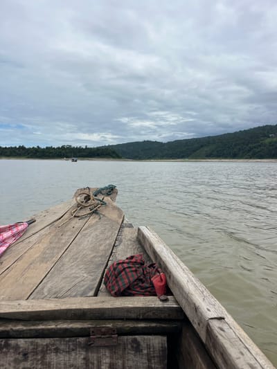 The image captures a view from the front of a wooden boat navigating a wide, calm body of water, likely the confluence of rivers in Jaflong, Bangladesh. The sky is overcast with grey clouds, suggesting a diffused light. The water is a muted, murky green, with gentle ripples on the surface. On the left side of the boat's bow, a pink and white umbrella is partially visible, laid flat. Two ropes, one thicker and natural-colored, the other thinner and greenish, are coiled on the wooden planks of the boat's deck. A folded piece of red and black plaid fabric rests on the deck towards the right. In the distance, a tree-lined shore forms a green, undulating silhouette against the horizon, with hills in the background. A small boat with a canopy is visible further out on the water. The foreground is dominated by the weathered, wooden structure of the boat, revealing planks, nails, and a metal hinge.