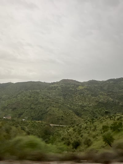 The image depicts a hillside landscape under an overcast sky, likely in or near Casabermeja, Spain. The terrain is primarily green with vegetation. The perspective suggests the photo was taken from a moving vehicle or a slightly elevated position. A hint of a roadway or built structure is visible further down the hillside, indicating some level of development in the area. The scene is muted and subdued, likely due to the weather conditions.