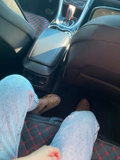 The image shows the interior of a car, likely in Herasymivka, Ukraine. The perspective is from inside the car, looking down at the passenger's legs and feet. The subject is a person wearing light-wash denim jeans and tan slip-on sandals. Their legs are crossed, with one foot resting on a black floor mat with red diamond-shaped stitching. The other foot is partially visible, also on a similar mat.

In the background, parts of the car's interior are visible, including a black leather car seat, a center console with a gear shift, and air vents. A red and black fabric accessory with the word "SPORT" printed on it is attached to the gear shifter. The lighting suggests it is daytime, with sunlight casting shadows inside the vehicle. The scene appears to be a mundane moment of someone sitting in a parked car. No specific emotions are discernible from the limited view of the subject.