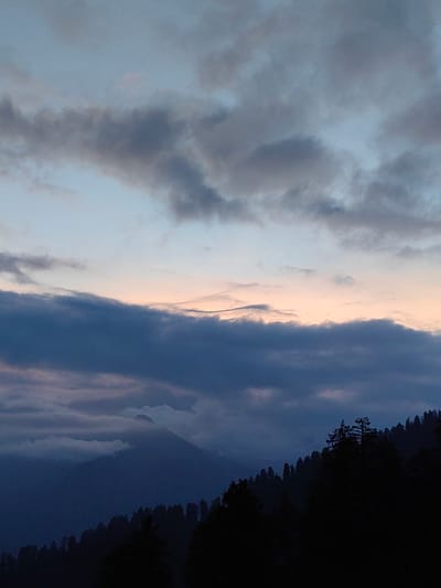 This is a vertical, medium-wide shot of a mountainous landscape near Aleo, India, at dusk. The sky dominates the upper two-thirds of the image, filled with layers of dark gray and blue clouds. A faint band of pale orange and pink light is visible on the horizon, suggesting the sun has recently set. The lower third of the image shows a silhouette of a dense forest of pine trees covering a steep mountainside. The trees are dark and indistinct, blending into the shadows. In the background, the outlines of more mountains are visible, shrouded in mist and fading into the darker sky. The overall mood is serene and somewhat dramatic, characteristic of twilight in a mountainous region.