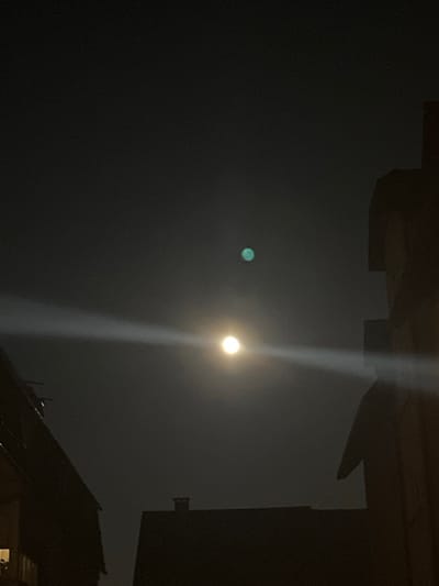 The scene is a nighttime urban environment, presumably in Tuttlingen, Germany. A dark sky dominates the upper frame. A prominent, circular, yellowish-white light source, consistent with the moon, is centered in the lower-middle portion of the frame. Positioned directly above and slightly right of this light is a smaller, distinct, circular, greenish-blue light. A horizontal, linear light artifact extends across the image, intersecting the primary light source. The lower and side portions of the image are framed by dark silhouettes of architectural structures. On the left, a building silhouette shows a gabled roofline with an antenna-like projection and a faint, rectangular light emission from a lower window. On the right, a darker, more angular building silhouette with varied roof levels occupies the frame's edge. A dark, rectangular chimney protrudes from a lower roofline silhouette visible between the primary structures in the background. No individuals or direct interactions are visible.