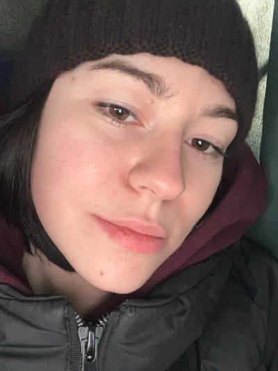 A close-up, slightly tilted portrait shows an individual, likely female, situated in Yurivka, Ukraine. The person wears a dark brown knitted beanie covering their upper forehead and dark brown, short hair is visible on the left side of the face. Their skin tone is fair with subtle redness on the cheeks and nose. The eyes are brown with minimal visible makeup, featuring natural, dark brown eyebrows. The lips are full and naturally colored, appearing slightly parted. A small mole is visible on the chin, and another minor blemish is present above the viewer's left eyebrow. The individual is dressed in a burgundy or dark red hooded sweatshirt beneath a black puffer jacket, identifiable by its visible silver zipper pull. The background is indistinct and dark, with a subtle greenish hue on the right, suggesting an out-of-focus interior environment. The subject maintains eye contact with the viewer, displaying a neutral to slightly somber expression.