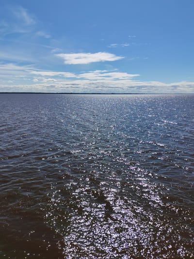 The image shows a body of water with a dark surface reflecting sunlight, under a blue sky with scattered white clouds. The horizon line is visible in the distance, with a thin strip of land along it. The water is rippled, and the sunlight creates a sparkling effect across the surface. The photo appears to be taken at a location with an expansive view over a large body of water. The image does not depict any features that would identify the location as Veregonets, Russia. There are no people visible.