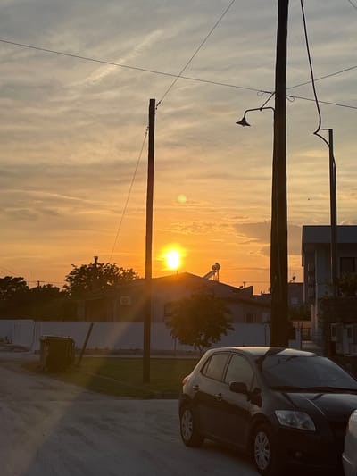 The image depicts an outdoor scene at dusk, likely in Chaidefto, Greece. The sun is low on the horizon, casting a warm, golden glow across the sky, with scattered clouds catching the light. The sky transitions from a pale blue-grey at the top to a vibrant orange and yellow near the sun.

Dominating the foreground are two utility poles with multiple wires strung between them, typical of an urban or suburban environment. One pole has an old-fashioned street lamp attached to it. In the background, buildings and trees are silhouetted against the sunset. A dark-colored car is parked on the right side of the frame, partially obscuring a building. The ground appears to be a paved surface, possibly a road or parking lot.

The overall mood is serene and peaceful, characteristic of a sunset. The low angle of the sun suggests it is late afternoon or early evening. No people are visible in the image. There is no discernible text in the image.