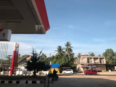 This image captures a vibrant street scene at a Pertamina gas station in Kutalawah, Indonesia, under a bright, clear blue sky, suggesting it is either morning or late afternoon.
In the foreground, the red and white canopy of the gas station is visible, along with a prominent "1" on a gas pump island. A tall Pertamina sign stands to the left, displaying "PERTAMINA", a number "13245410", and a list of fuel types: "PERTAMAX", "PERTALITE", "PREMIUN" (likely Premium), "BIO SOLAR", and "DEX". Below this, a partially obscured sign reads "MOHON MAAF SEDANG WAKTU SHOLAT", which translates to "Apologies, during prayer time".
The midground and background depict a lively street. A person is visible near a yellow truck covered with a blue tarp, possibly attending to a scooter. Other vehicles on the road include a white car and a red minivan, the latter appearing to be in motion. The area is lined with various buildings: a house with a distinctive roof, an unfinished multi-story concrete structure, and other smaller establishments. One small sign is visible, reading "Bak Kopi". Abundant tropical foliage, including numerous tall palm trees and a conifer-like tree, dots the landscape, contributing to the warm, tropical setting. The scene portrays a typical daily activity in a bustling roadside area.