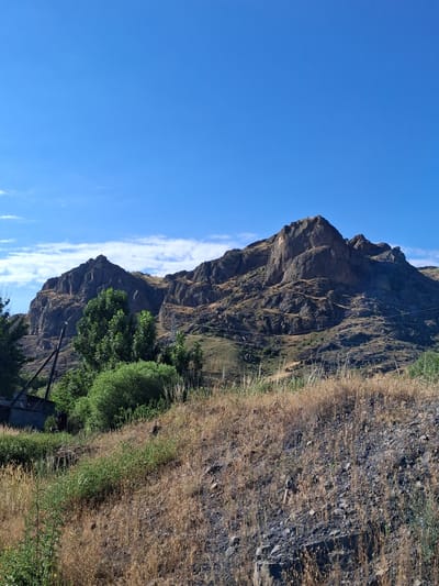 The image shows a rocky mountain landscape under a clear blue sky with some wispy clouds. In the foreground, there is dry, yellowed grass and some scattered rocks. Behind the dry grass, there are patches of green bushes and trees. A dark, angular structure, possibly a building or shed, is partially visible on the left side, near some power lines and poles. The main subject of the image is the imposing, rugged mountain range that dominates the background. The rocks of the mountain are dark and textured, with visible crevices and sharp angles, suggesting a natural, eroded formation. The overall impression is a rural, natural setting. This scene is located in the vicinity of Malishka, Armenia, characterized by such mountainous terrain.