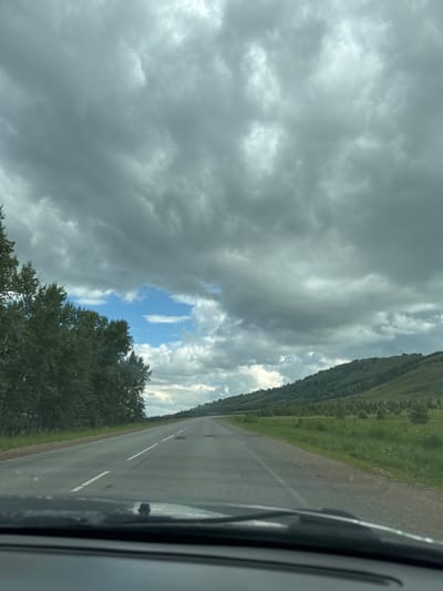 The image depicts a view from inside a vehicle driving on a two-lane asphalt road. The sky is filled with dramatic, dark gray clouds, with a small patch of blue sky visible in the distance. To the left of the road, a dense line of green trees is present. To the right, rolling hills covered in green grass and scattered trees recede into the distance. The road itself is marked with white lines, indicating lanes and traffic direction. The foreground shows the dashboard and windshield wipers of the vehicle. The scene appears to be a rural or semi-rural landscape, possibly on the outskirts of a city, and it is not possible to determine the specific location within the city of Uralsk, Russia from this image alone. There are no people or animals visible in the image.