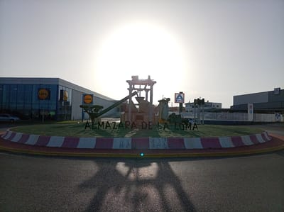 The image shows a roundabout in Albox, Spain, featuring a large metal sculpture in the center. The sculpture appears to be an industrial or agricultural machine, possibly a replica of an olive oil press, given the context of "Almazara de la Loma" which translates to "Olive Mill of the Hill." The roundabout has a grassy area with the name of the place spelled out in large metal letters. The roundabout's edge is lined with red and white striped barriers.

Surrounding the roundabout are several commercial buildings. To the left, a modern building with large glass windows is visible, with prominent "Lidl" logos. To the right, another building with a "Lidl" logo is present, as well as a building with an "ALDI" sign. Further to the right, a parking sign is visible. The sun is high in the sky, creating a bright glare and casting long shadows across the roundabout and the road. The road surface is asphalt. There are no people clearly visible in the image, though silhouettes can be seen inside the Lidl building. The overall environment suggests a commercial area on the outskirts of the town.