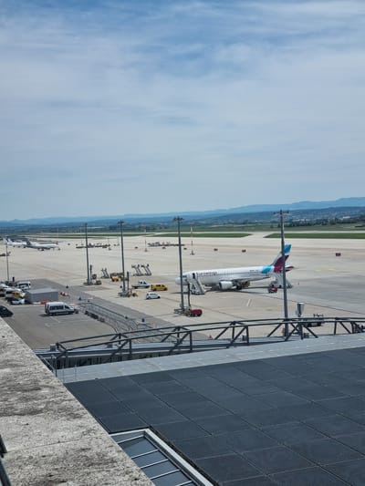 The image shows an airport scene in Echterdingen, Germany. An Eurowings airplane, identifiable by its teal and purple tail, is parked at a gate. A jet bridge is connected to the plane. Various service vehicles are positioned around the aircraft on the tarmac. Light poles are visible along the edge of the tarmac, and in the background, a line of trees and distant hills are present. A portion of the airport terminal building is visible on the right side of the image. The sky is bright blue with scattered clouds.