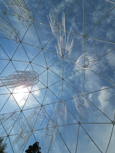 The image shows a view looking up through the geodesic dome structure of the Ulitkino Aviary, located in Ulitkino, Russia. The dome is composed of white metal struts forming triangular panels. Hanging within the dome are several decorative elements that resemble jellyfish or chandeliers. These decorations consist of circular frames from which numerous thin, transparent strands hang down, creating a cascading effect. The sky, a clear blue with scattered white clouds, is visible through the dome's panels. The sun is also visible, shining brightly and causing lens flare. In the lower portion of the image, a dark green tree is visible outside the dome structure.