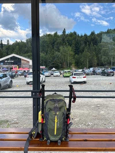The image depicts a backpack and two trekking poles resting on a wooden bench in Szczyrk, Poland. In the background, a parking lot is filled with various cars, and beyond that, a dense forest covers the hillside. A building with a prominent "SZCZYRK" logo and "Infocentrum" signage is visible on the left side of the frame, suggesting a tourist or outdoor activity hub. The sky above is partly cloudy, with a mix of blue sky and white clouds. The scene appears to be a rest stop or waiting area, likely at a trailhead or near an outdoor attraction in the mountainous region of Szczyrk.