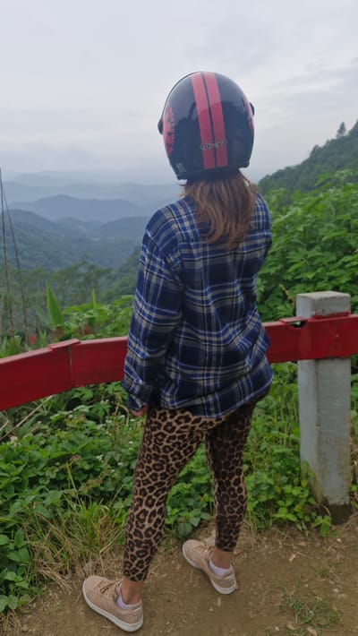 A person stands with their back to the camera, facing a panoramic view of lush, mist-shrouded mountains. They are wearing a dark blue and yellow plaid shirt, leopard-print leggings, and tan sneakers. On their head, they wear a black helmet with two red stripes and the word "AVEX" printed on it. A red railing, partially obscured by greenery, runs horizontally in the foreground. The ground is a mix of dirt and grass. The location is identified as Samoeng Tai, Thailand, suggesting a scenic mountainous area.