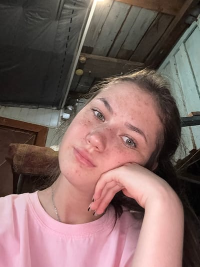 The image is a close-up selfie of a young woman with freckles, looking directly at the camera. She has light brown hair and is wearing a pink t-shirt and a silver chain necklace. Her right hand is propping up her head, and her left hand is resting on her chest. The background appears to be an indoor setting with wooden walls and ceiling. The lighting is dim and likely artificial. The location provided is the city of Rezh, Russia.