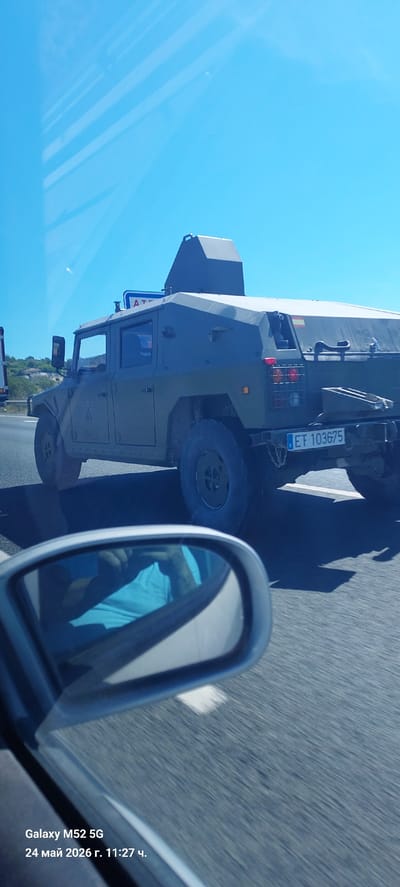 The image shows a military vehicle with license plate "EF 103675" on a highway. The photograph appears to have been taken from the passenger seat of a car, evidenced by the car's side mirror visible in the foreground. The environment is sunny with a clear blue sky. The location is likely in Rotglà i Corberà, Spain, based on the license plate, although there is no definitive visual evidence to confirm this beyond the license plate's association with the country. The image also contains a timestamp "Galaxy M52 5G 24 Май 2026 г. 11:27 ч."