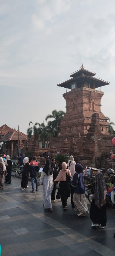 The image shows a group of people walking on a paved area in front of a large brick clock tower in Kauman, Indonesia. The clock tower has a square base and a tiered roof with decorative elements. A clock face is visible on one side of the tower. Several palm trees are visible behind the tower. The people in the foreground are mostly wearing headscarves and modest clothing. Some are walking towards the tower, while others are walking away. The ground is paved with large, dark gray square tiles. The sky is overcast. There are also some motorcycles and a shopping cart visible on the right side of the image. The architecture of the buildings in the background suggests a traditional Indonesian style.