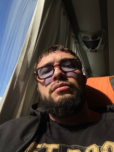 The image shows a close-up, low-angle, selfie-style shot of a man with a beard, wearing sunglasses and a black zip-up jacket over a black t-shirt with a gold graphic design. He is seated on an orange and grey seat, likely in a vehicle such as a bus, as suggested by the window and curtains on the left and the overhead interior on the right. The man's face is illuminated by sunlight streaming through the window. The background shows a blue sky with some clouds and the interior of the vehicle. The location is specified as Glukhovo, Russia.