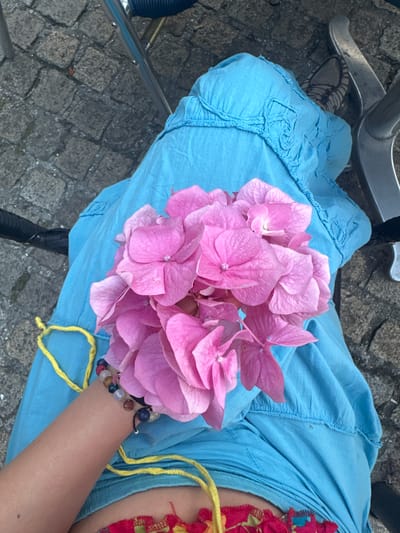 A person wearing a blue dress and a colorful garment at the waist, sits on a chair outdoors, holding a bouquet of pink hydrangeas. The bouquet is tied with a yellow string. The person's arm has a beaded bracelet on their wrist. The ground is paved with cobblestones. The location is in the city of Noyon, France.