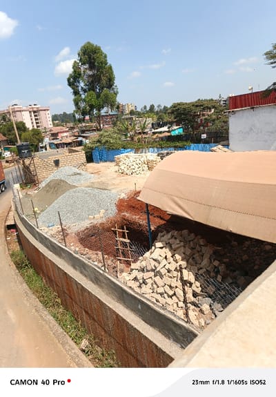 The image captures an urban construction site. Piles of gravel and building stones rest near an excavated area accessed by a wooden ladder. A tan-colored tarp provides partial cover. Beyond the site, a bustling cityscape with buildings and trees stretches into the distance under a bright sky with scattered clouds. A barrier wall separates the construction zone from the adjacent road. The scene is captured with a smartphone camera, as indicated by the watermark.