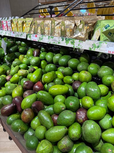 The image captures a vibrant display of produce and packaged snacks in a grocery store, likely in Kelapa Dua, Indonesia. The scene is dominated by a large, overflowing bin of fresh avocados, which occupy the right and bottom portions of the frame. Most avocados are a rich green color, indicating various stages of ripeness, while some display darker, purplish-brown hues. Several avocados feature small white stickers with the text "agp fresh."

To the left, a metal display rack holds numerous bunches of ripe yellow bananas, arranged vertically. Adjacent to the bananas, an upright shelf showcases an array of packaged freeze-dried fruit snacks. Prominently featured are multiple gold-colored pouches of "DURIAN FREEZE DRIED" snacks, with some showing Chinese characters "有零食" (meaning "have snacks") and the brand "IRSnack Jurs." These packages highlight "ALL NATURAL," "100% REAL FRUIT," "NO SUGAR ADDED," and "PRESERVATIVES FREE" claims. A price label for the durian snacks reads "69.800," indicating Rp 69,800. Further up the shelf, green packages from "Eat Me Brand" are visible, specifically "Baby Blond" "FREEZE DRIED MELON BITES," with a 15GR weight and a price of "Rp 34.80" (Rp 34,800). Other green packages labeled "MANGO" are also stacked below the durian snacks. The overall impression is a well-stocked fresh produce and snack section.
