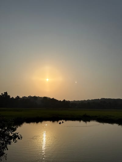 The image shows a sunset scene at a lake in Salvador du Mundu, India. The sun is positioned above a tree-lined horizon, with its reflection visible on the water's surface. Two small boats are visible on the lake, with the silhouette of a person in one of them. The sky fades from a pale grey to an orangish yellow color.