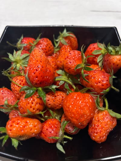 A high-angle, close-up shot showcases a black bowl filled with fresh strawberries. The strawberries are ripe and vibrantly red, with visible seeds dotting their surfaces. Green stems and leaves are attached to many of the fruits, suggesting they were recently picked. The lighting is bright, highlighting the glossy texture of the strawberries. The background is out of focus, but appears to be a light-colored, textured surface, possibly a tablecloth or countertop. The setting suggests a home kitchen or a place where fresh produce is prepared or displayed. No people are visible in the image, and there is no visible text. The image captures a simple yet appealing still life of fresh fruit.