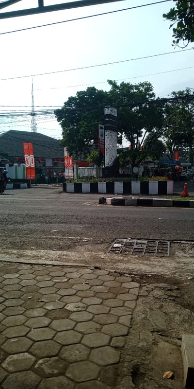 This daylight image portrays an urban street scene at a roundabout in Bojongherang, Indonesia. The foreground consists of hexagonal concrete pavers leading to an asphalt road, where a square metal drain cover is visible. The central roundabout island is edged by a black and white checkered curb. A tall, multi-faceted monument, primarily black and white with intricate patterns and some illegible text, stands prominently on the island, surrounded by green trees and foliage. Two large, vertical red banners are positioned behind the monument; one clearly displays "BO" and "00" alongside the partial word "BANK." A dark metal fence runs along the island's perimeter. In the background, a building with a dark, sloped roof is visible to the left, and a prominent lattice communication tower ascends against a light blue sky, intersected by numerous overhead power lines. A motorcycle is captured in motion on the left side of the asphalt road, and an orange traffic cone is present near the roundabout's edge on the far right.