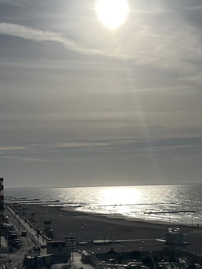 The image depicts a beach scene in Palavas-les-Flots, France, under bright, hazy sunlight. The sun, prominent in the sky, casts a strong glare on the water, creating a shimmering effect. The beach itself appears wide, with dark sand that may be wet from the tide. The coastline is lined with structures that appear to be buildings and possibly lifeguard stations. A promenade runs along the beach, with street lights and railings visible. The sky is heavily clouded, obscuring a clear view of the horizon.