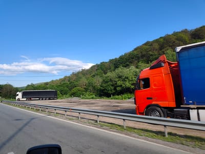 The image captures a roadside scene in Gorno Kamartsi, Bulgaria, on a clear, sunny day. The foreground shows a paved road with a metal guardrail running along its edge. Beyond the guardrail, there is a flat, open area with some patches of gravel and puddles, bordered by a lush green hillside covered in trees. Two large trucks are visible. In the distance on the left, a white truck with a black trailer is parked. Closer to the right side of the image and in motion, an orange truck with a blue trailer is passing by. The sky is a vibrant blue with a few wispy clouds. The overall impression is one of a rural or semi-rural transportation hub.