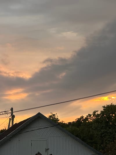The image captures an outdoor scene at dusk. The sky is filled with a mix of dark gray and wispy clouds, with areas of bright orange and yellow light indicating the setting sun peeking through. Silhouetted trees frame the right side of the image, their dark green foliage contrasting with the illuminated sky. In the foreground, the white, corrugated metal siding of a building is visible, with a sloped roofline. Power lines and a utility pole are also present, strung across the sky, suggesting a semi-rural or suburban setting. The overall mood is somewhat dramatic due to the stormy-looking clouds and the intense sunset colors. There is no visible text in the image.