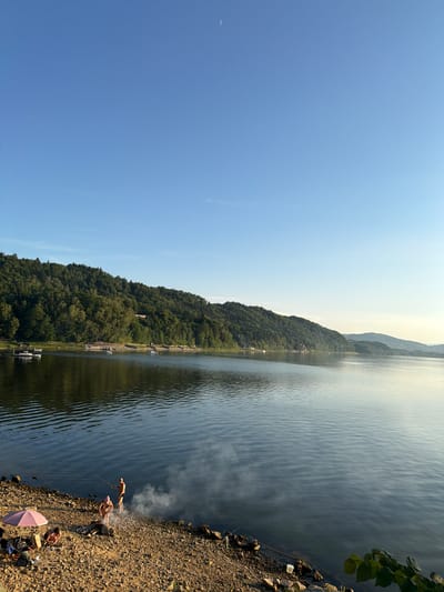 The image shows a serene landscape at the lake in Lipie, Poland. The clear blue sky is vast, with a faint crescent moon visible in the upper portion. A dense, verdant forest covers a rolling hill that slopes down to the water's edge. The calm lake's surface reflects the surrounding greenery and the sky.
In the foreground, a rocky shore is dotted with a few individuals engaged in recreational activities. Two people are standing near the water's edge, one appears to be fishing with a rod. A small campfire is burning, emitting a gentle plume of smoke. To the left, another person is seated on the shore, with a pink umbrella providing shade. Camping gear, including bags and a bicycle, are also visible on the shore. The overall scene depicts a tranquil day at the lake.
