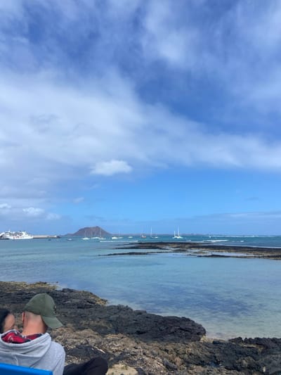 The image captures a view from a rocky shoreline in Corralejo, Fuerteventura, Spain (latitude: 28.737, longitude: -13.867). The scene features a vast expanse of blue water stretching towards the horizon, where several boats are anchored. The water is clear and turquoise, with a rocky seabed visible beneath the surface. In the distance, a mountainous landmass emerges from the sea. The sky is a vibrant blue, dotted with white, fluffy clouds. On the rocky shoreline, a lone figure sits with their back to the camera, wearing a green baseball cap and a blue hooded sweatshirt over a plaid shirt. The person appears to be gazing at the boats and the distant landmass. The scene portrays a peaceful and serene setting, offering a picturesque view of the coastal landscape.